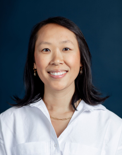 A woman with straight black hair wearing a white collared shirt and gold jewelry stands in front of a dark blue background, smiling at the camera.